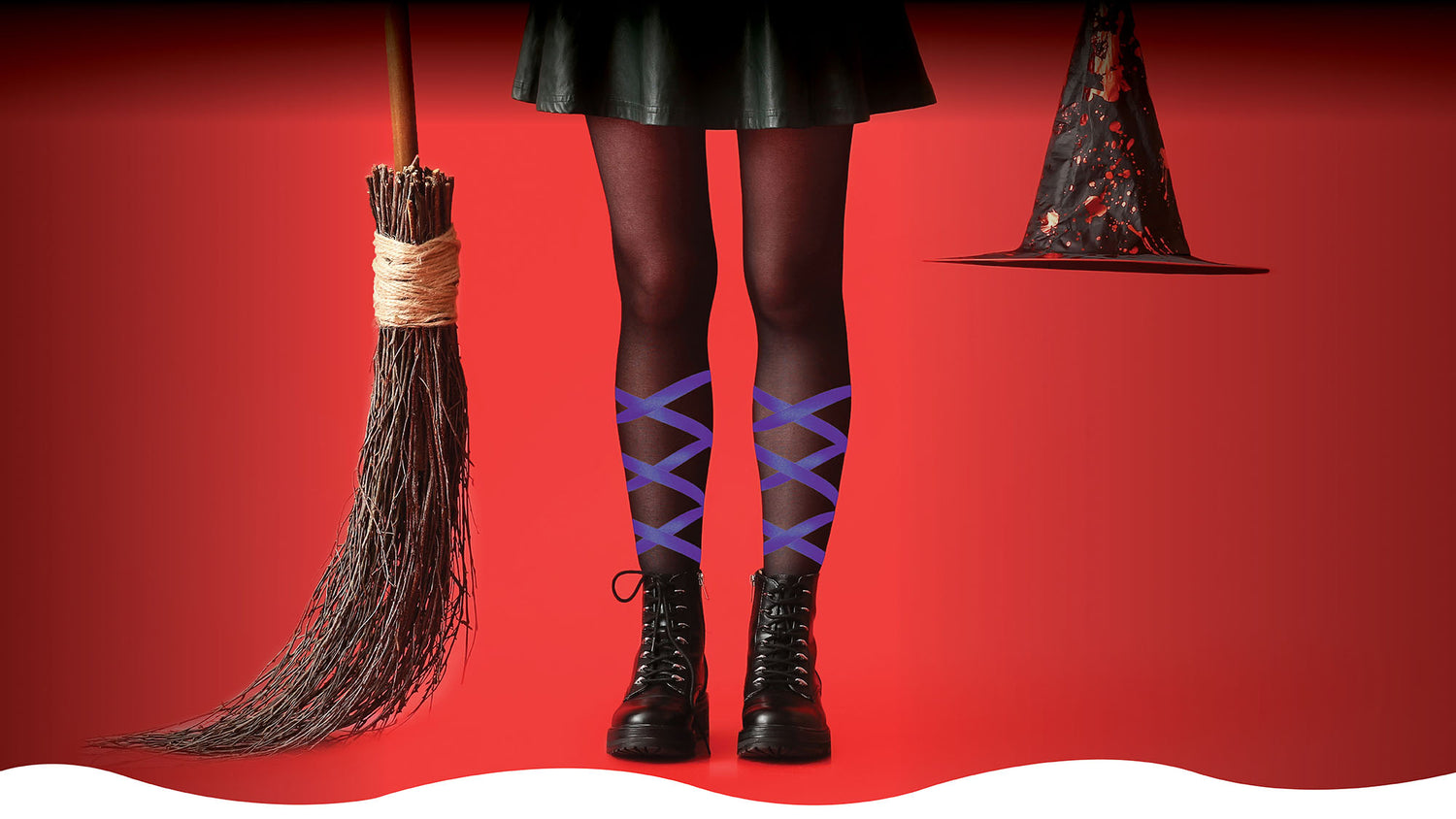 Person wearing black skirt, tights with purple Solid LegUps and black boots on a red background with a broom and witch hat.